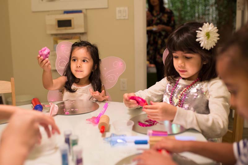 Two young girls in fairy accoutrements including fake mesh wings, doing crafts with colored gel at a white table