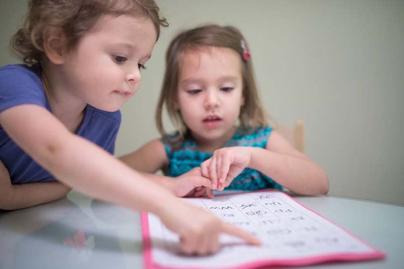 Two young girls working together on learning the alphabet. One girl is pointing at a worksheet on a table they are sitting at.