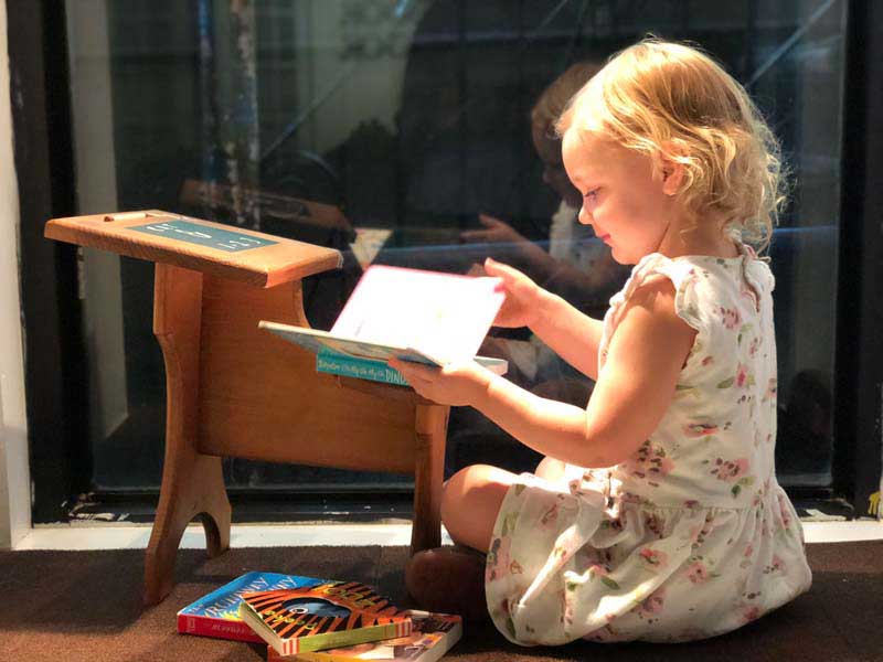 Young girl in a floral fress reading at a small desk in the sunlight