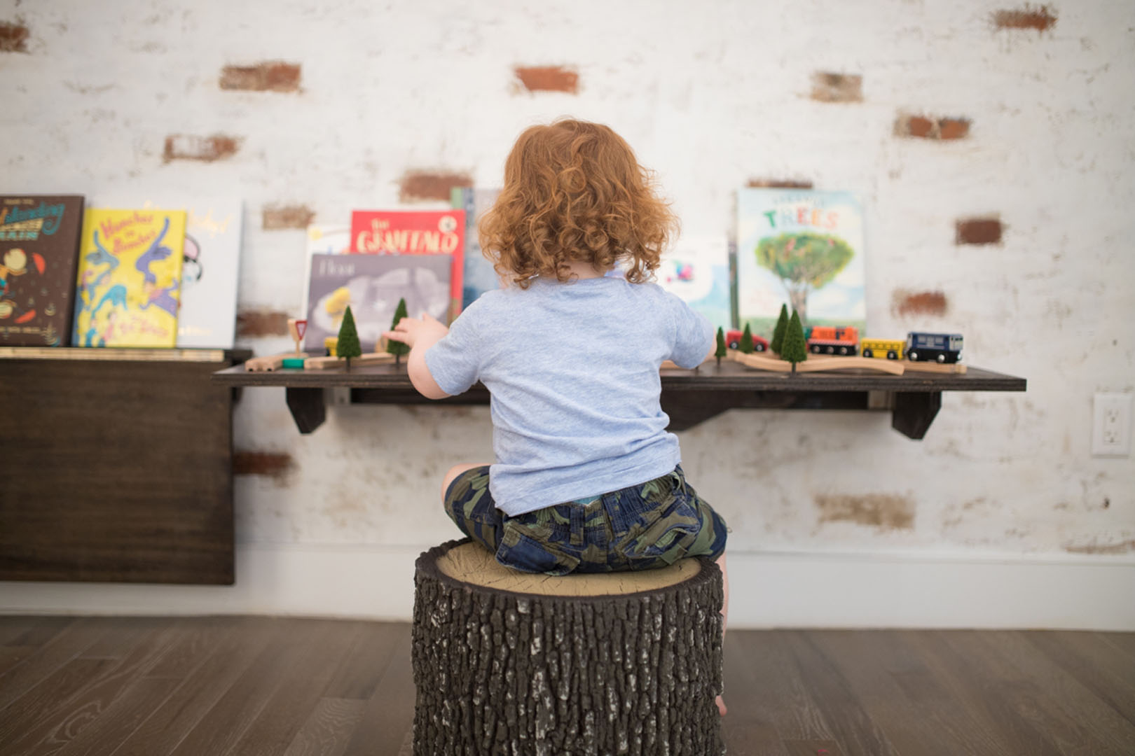 Little girl sitting on a standing log as a chair at a wall desk, the wall is decorated with childrens books and toys