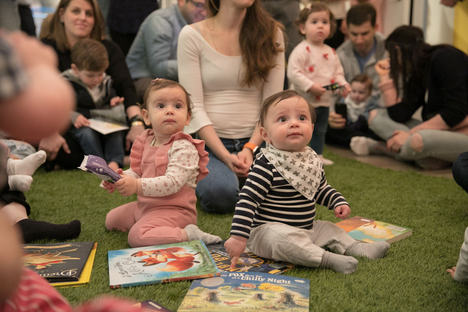 Two toddlers sitting on the floor with books, looking up at a teacher reading to them. Their mothers are sitting behind them.