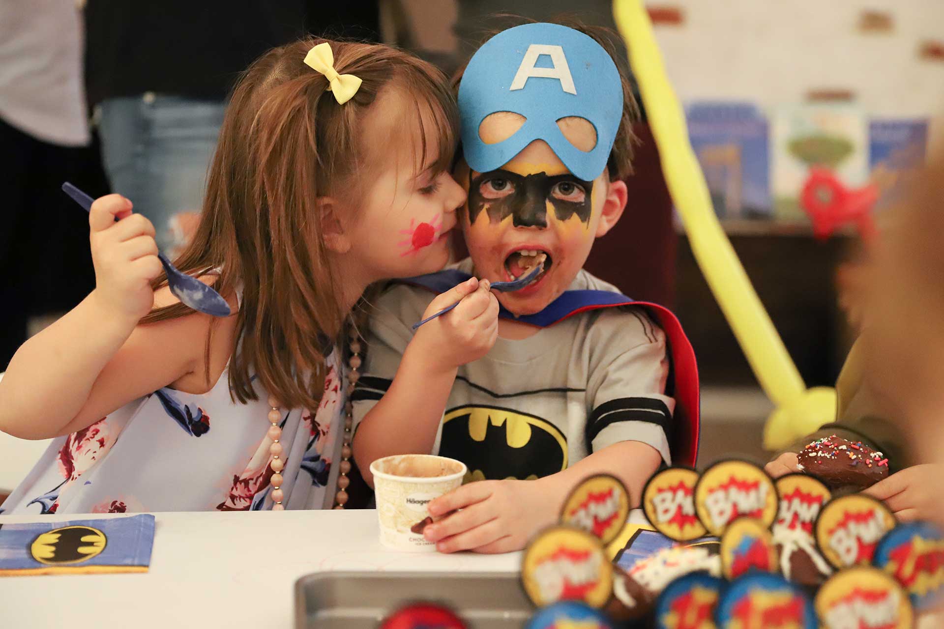 A boy and a girl wearing clown makeup and party clothes eating a dessert while the girl gives the boy a kiss on the cheek