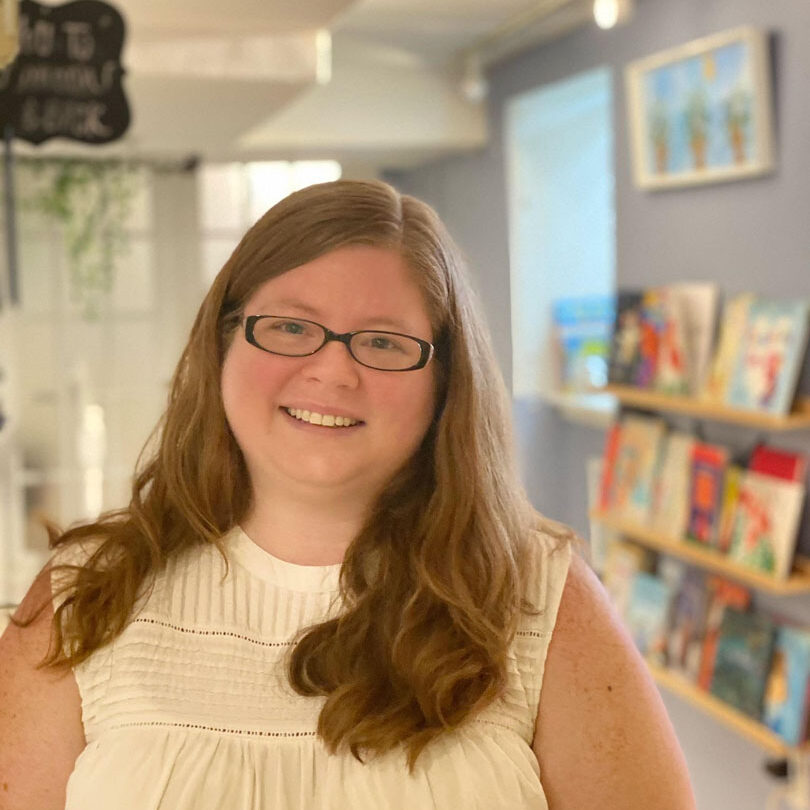 Heather Jenkins standing in Book Nook with a wall of books in the background. She is wearing glasses and smiling in a white tank top shirt.