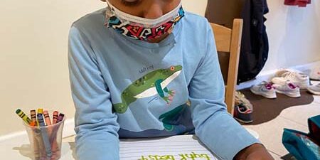 Young girl wearing a mask and shirt with turtles on it working on alphabet learning worksheets