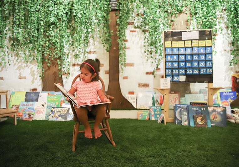 Young girl reading a book in a classroom with astroturf and trees crafted on the walls